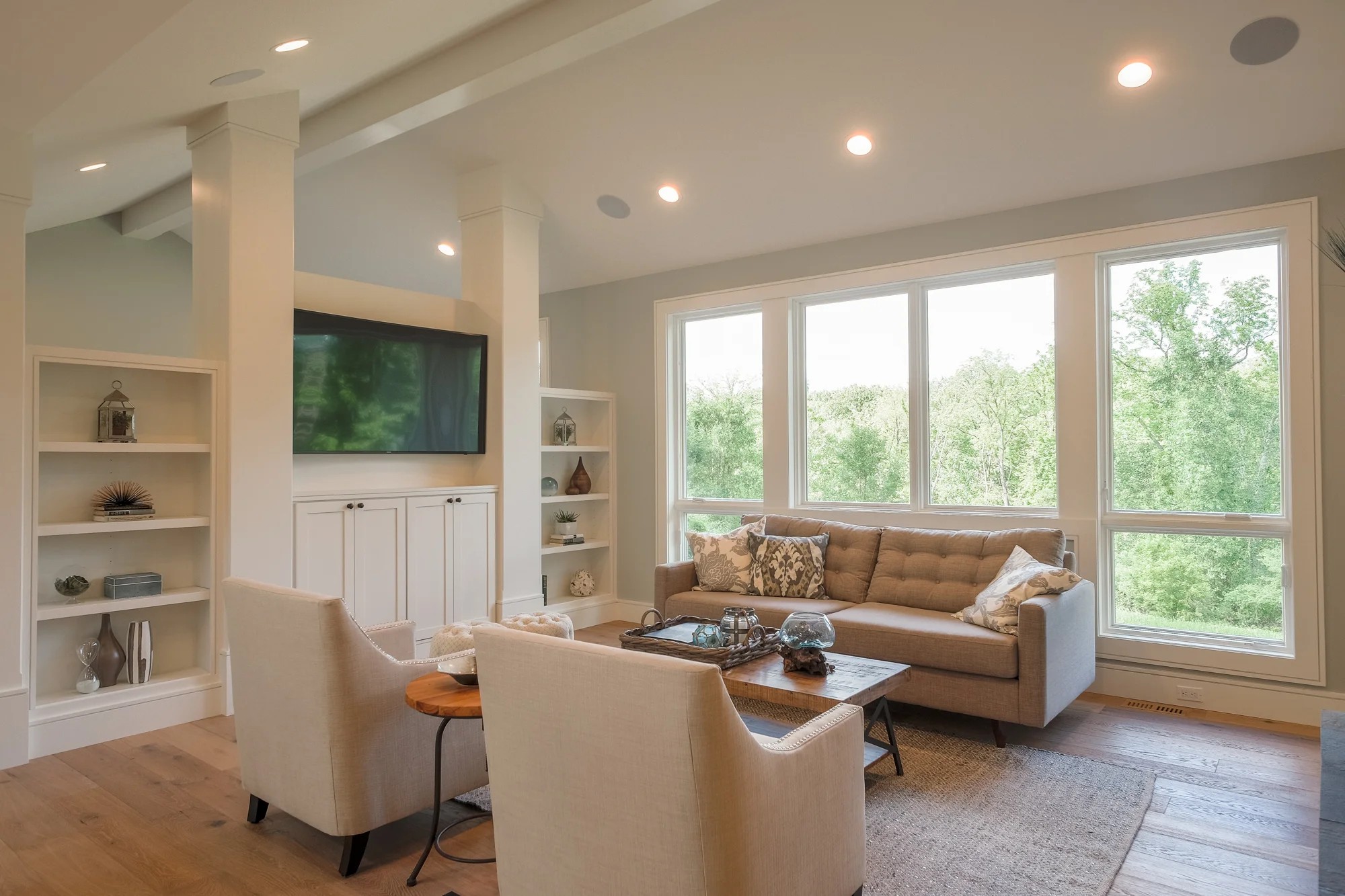 Living room with white built-ins flanking the television, a tan sofa, a vaulted ceiling, and a wall of windows to the woods.