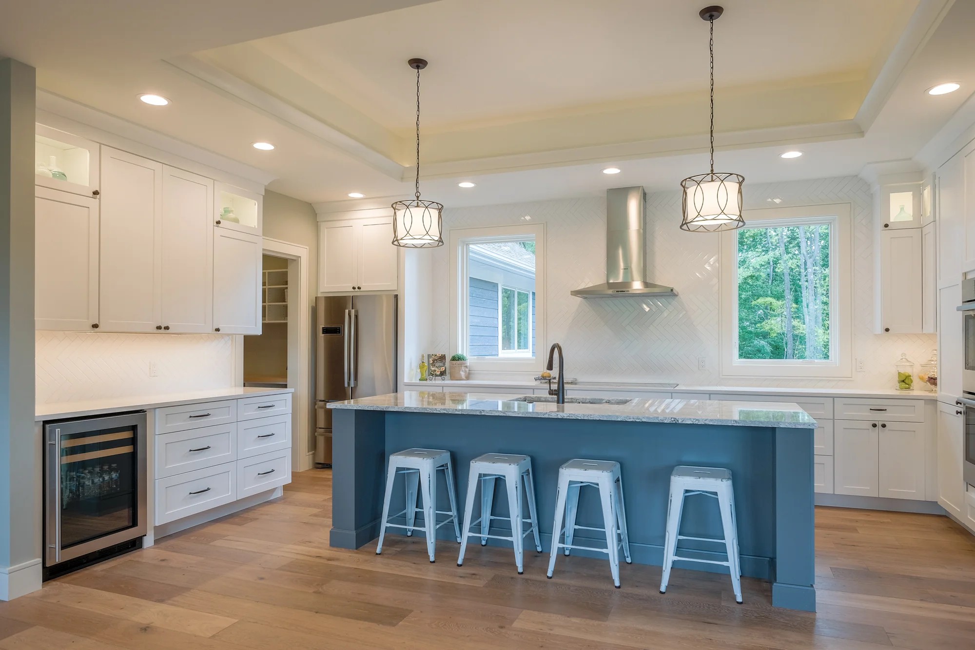 Wide kitchen view — white shaker cabinets, a navy island with marble counters, a beverage fridge, a stainless hood, and pendant lights.