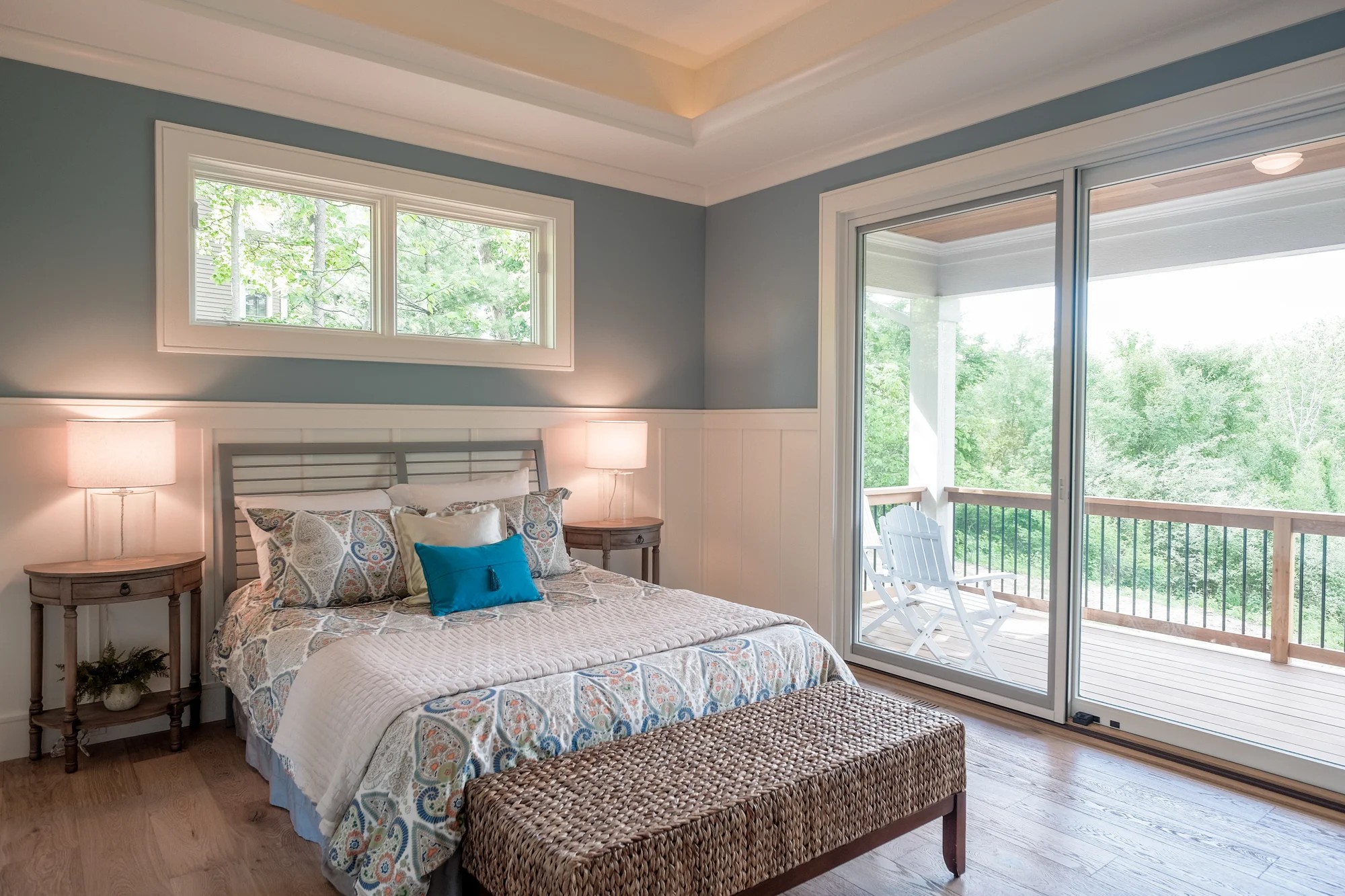 Primary bedroom with blue-gray walls, board-and-batten wainscot, a tray ceiling, and sliding doors opening to a covered deck overlooking the trees.