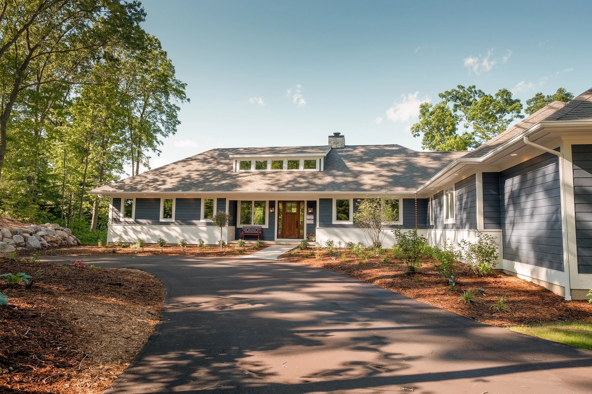 Fairbanks Estates — front three-quarter exterior with gray-navy siding, a gabled center entry, and a curved drive through a wooded lot.